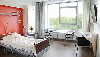 Helles Patient:innenzimmer mit Pflegebett, Tisch mit zwei Stühlen, Schreibtisch, Fernseher und großem Fenster.