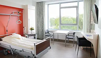 Helles Patient:innenzimmer mit Pflegebett, Tisch mit zwei Stühlen, Schreibtisch, Fernseher und großem Fenster.