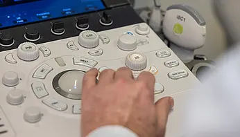 Eine Hand bedient die Tasten eines Ultraschallgeräts. Auf dem Gerät sind verschiedene Einstellknöpfe und Bedienelemente zu sehen.