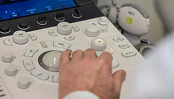 Eine Hand bedient die Tasten eines Ultraschallgeräts. Auf dem Gerät sind verschiedene Einstellknöpfe und Bedienelemente zu sehen.