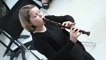 Person sitzt auf einem Stuhl und spielt eine Blockflöte; Notenständer und weitere Stühle sind im Hintergrund zu sehen.