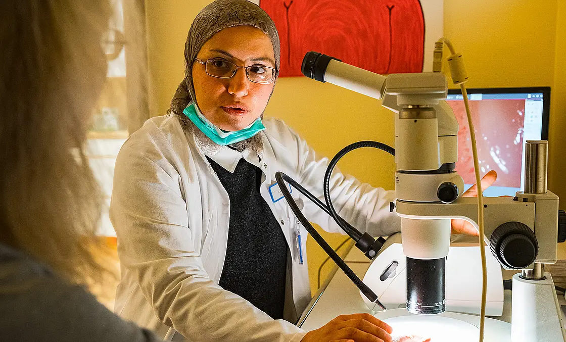 Behandelnde Person erklärt eine Untersuchung an einem Mikroskop, während eine Patientin ihre Hand auf die Arbeitsfläche legt.