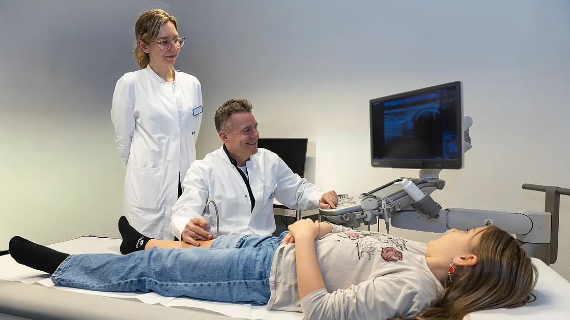 Dr. PD Daniel Windschall sitzt neben einer Untersuchungsliege und führt bei einem liegenden Kind eine Ultraschalluntersuchung durch. Eine weitere Ärztin steht daneben und schaut auf den Monitor.