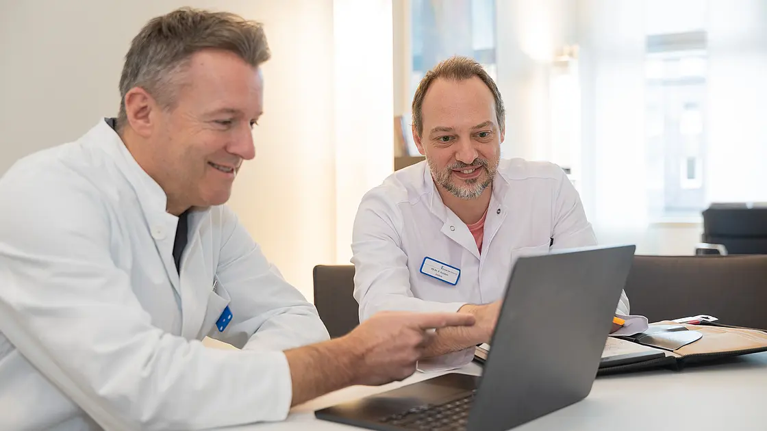 PD Dr. Daniel Windschall und PD Dr. Peter Korsten sitzen an einem Tisch und betrachten gemeinsam Inhalte auf einem Laptop.