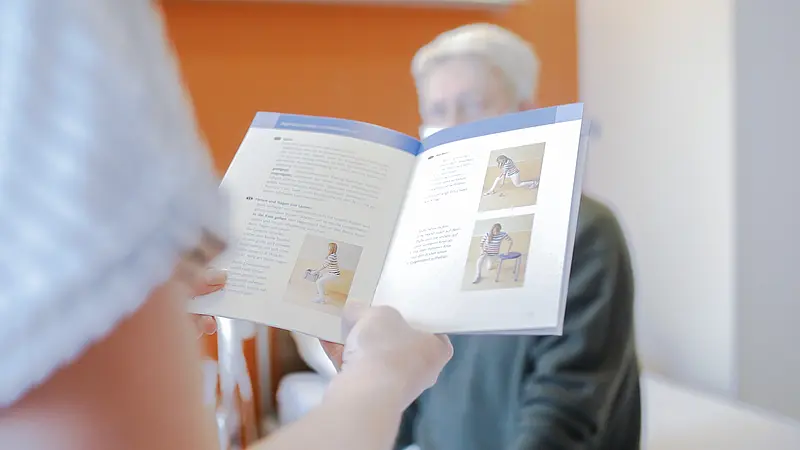 Eine Person hält eine Broschüre mit Übungsanleitungen für den Rücken, im Hintergrund sitzt eine weitere Person.