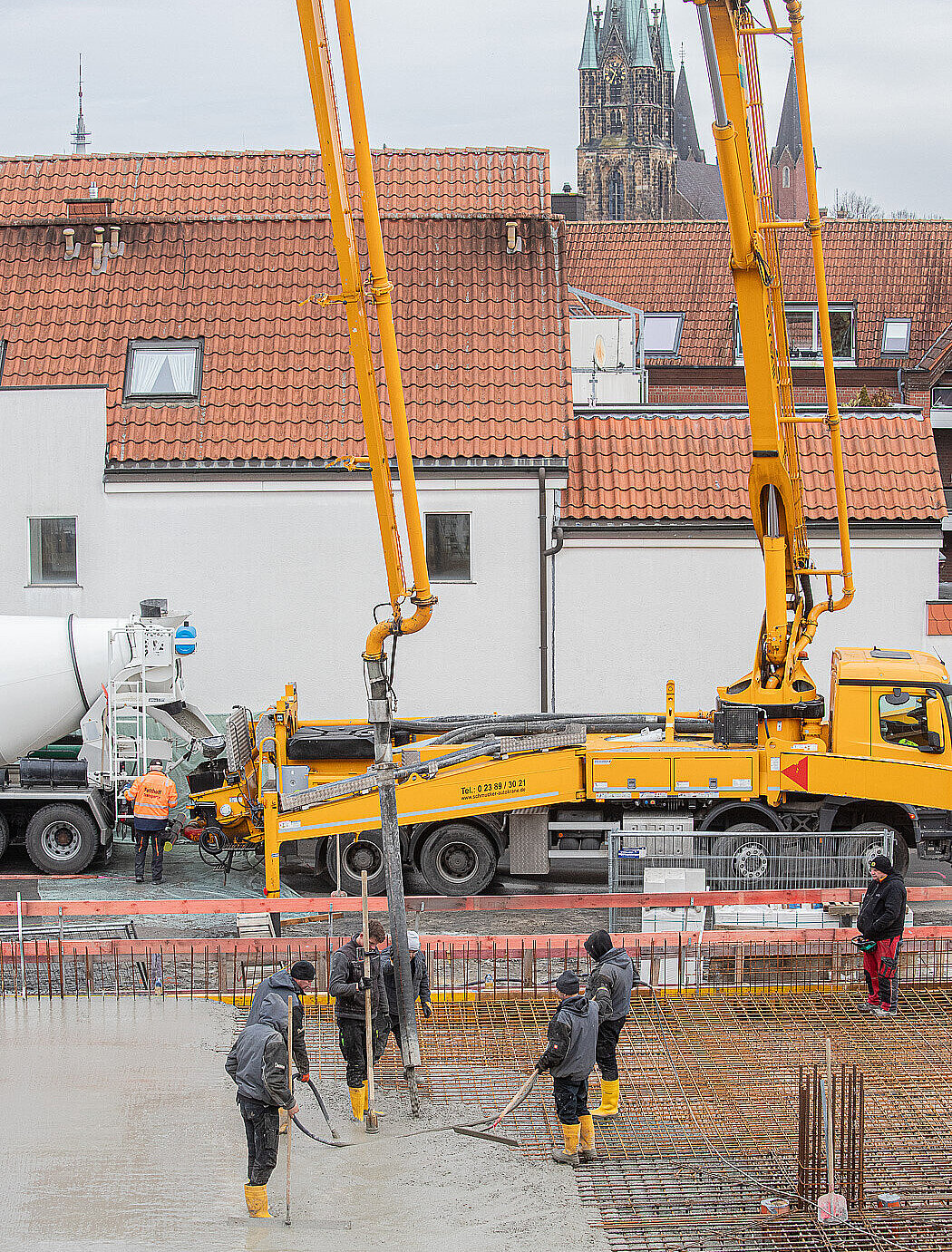 Ein Kran steht vor einer Baustelle, daneben ist ein Betonmischer. Auf der Baustelle stehen Bauarbeiter, die Beton gießen.