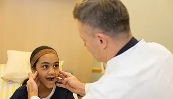 PD Dr. Daniel Windschall untersucht das Gesicht eines sitzenden Kindes und legt dabei seine Hände an die Wangen. Das Kind schaut ihn an und hat den Mund geöffnet.