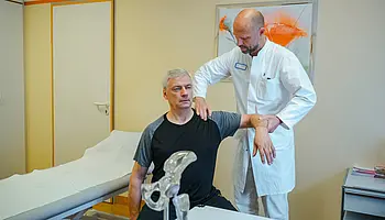 Ein Arzt prüft die Beweglichkeit der Schulter eines sitzenden Patienten in einem Untersuchungszimmer.