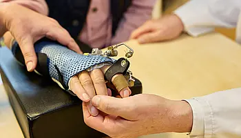 Mehrere Hände justieren gemeinsam eine Handorthese auf einer Ablage, konzentriert auf die funktionelle Einstellung.