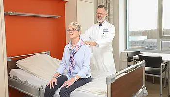 Chefarzt Dr. Christian Brinkmann legt einer sitzenden Patientin die Hände auf die Schultern in einem Patientenzimmer.