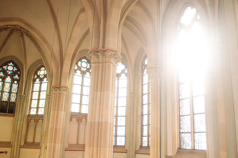 Blick in den Innenraum einer Kapelle mit hohen Säulen, Spitzbögen und großen Buntglasfenstern; Tageslicht fällt durch die Fenster in den Raum.