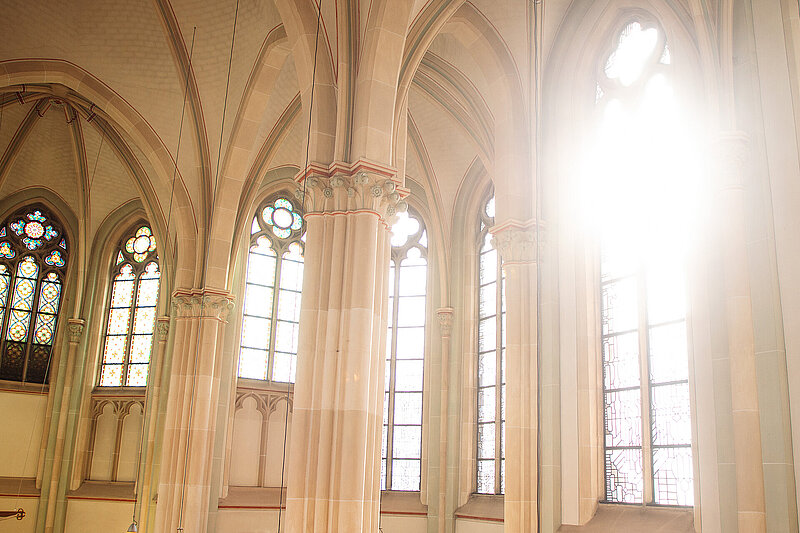 Blick in den Innenraum einer Kapelle mit hohen Säulen, Spitzbögen und großen Buntglasfenstern; Tageslicht fällt durch die Fenster in den Raum.
