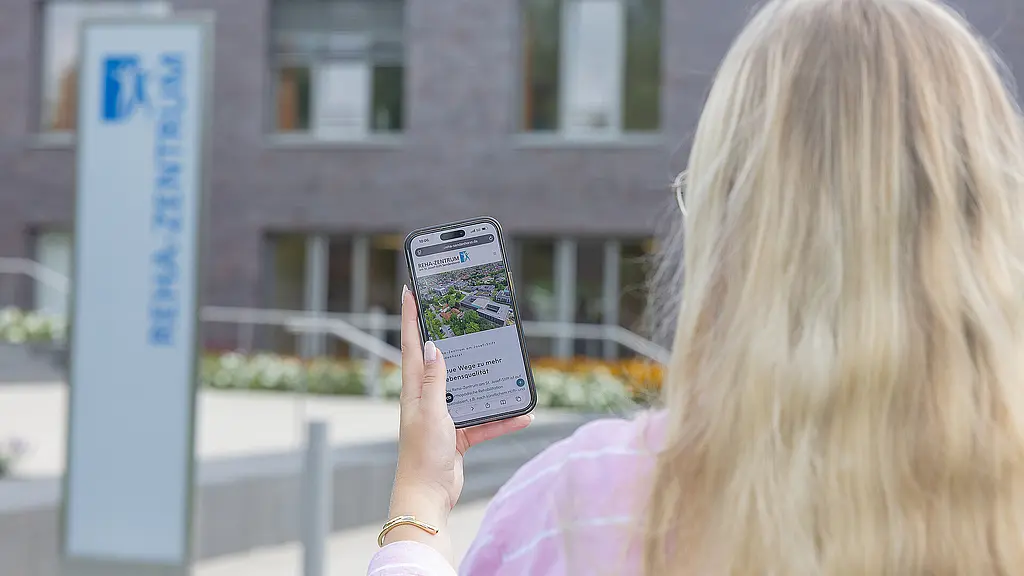 Ein Foto von einer Auszubildenden, die vor dem Reha-Zentrum steht. ie hat ein Handy in der Hand, auf dem die neue Reha-Website zu sehen ist.