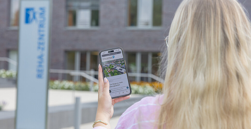 Ein Foto von einer Auszubildenden, die vor dem Reha-Zentrum steht. ie hat ein Handy in der Hand, auf dem die neue Reha-Website zu sehen ist.