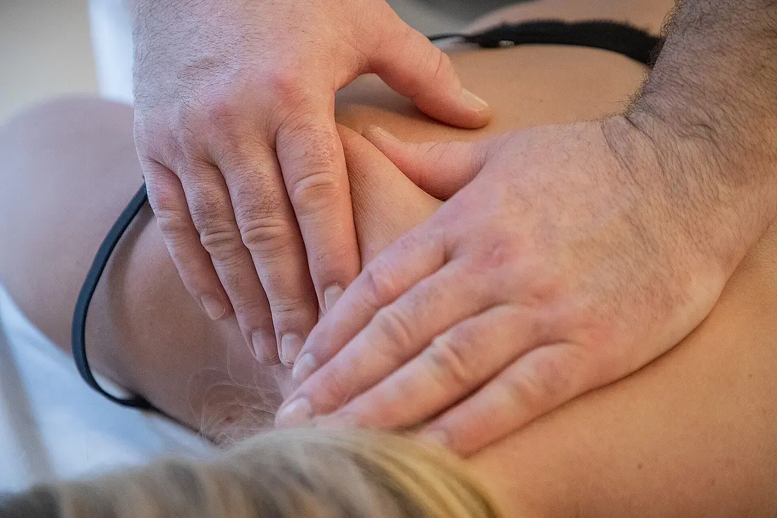 Ein Therapeut behandelt mit beiden Händen den Schulter- und Nackenbereich einer auf dem Bauch liegenden Person auf einer Therapieliege.