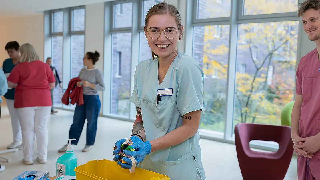Eine Mitarbeiterin in Arbeitskleidung und Handschuhen zeigt im Rahmen der Aktion Saubere Hände gesammelte Handschuhe an einem Informationsstand; im Hintergrund stehen weitere Personen im Klinikflur.