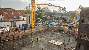 Großflächige Baugrube mit verlegter Bewehrung; ein Betonpumpenarm reicht über die Baustelle, während Beton in die vorbereitete Bodenplatte eingebracht wird. Mehrere Bauarbeitende stehen auf der Fläche.