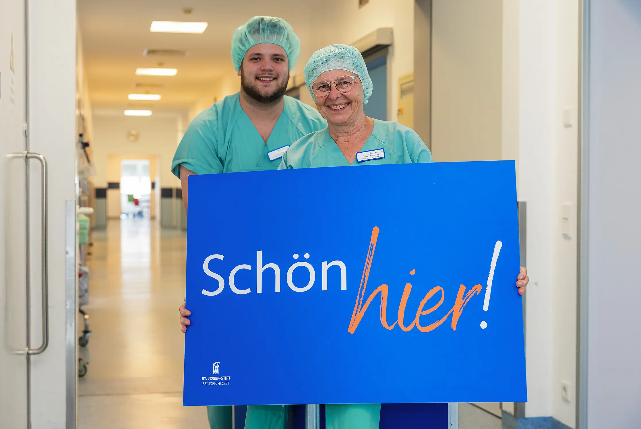 Zwei Mitarbeitende in OP-Kleidung stehen auf einem Flur und halten ein Schild mit der Aufschrift „Schön hier!“.
