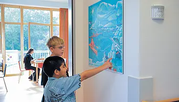 Zwei Kinder betrachten ein farbiges Kunstbild an der Wand eines Klinikflurs.