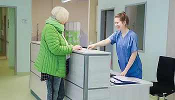 Eine Patientin steht im Empfangsbereich der Klinik für Ambulante Operationen & Sporttraumatologie und spricht mit einer Mitarbeiterin.