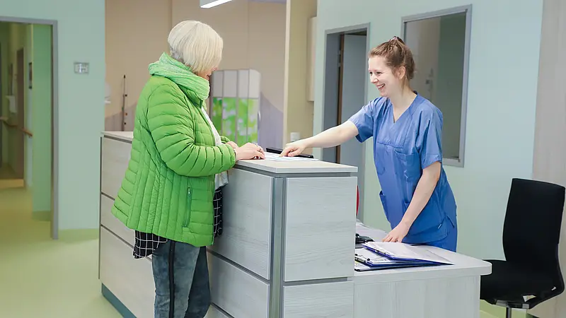 Eine Patientin steht im Empfangsbereich der Klinik für Ambulante Operationen & Sporttraumatologie und spricht mit einer Mitarbeiterin.