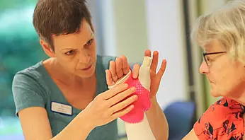 Eine Ergotherapeutin passt eine pinke Handschiene an der Hand einer Patientin an und überprüft die Fingerstellung.