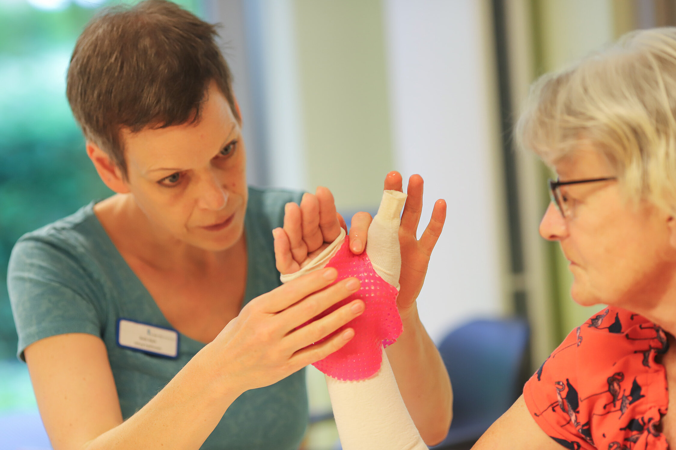 Eine Ergotherapeutin passt eine pinke Handschiene an der Hand einer Patientin an und überprüft die Fingerstellung.