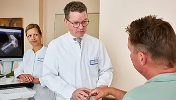 Dr. med. Ansgar Platte hält die Hand eines sitzenden Patienten und überprüft gezielt die Beweglichkeit einzelner Finger.