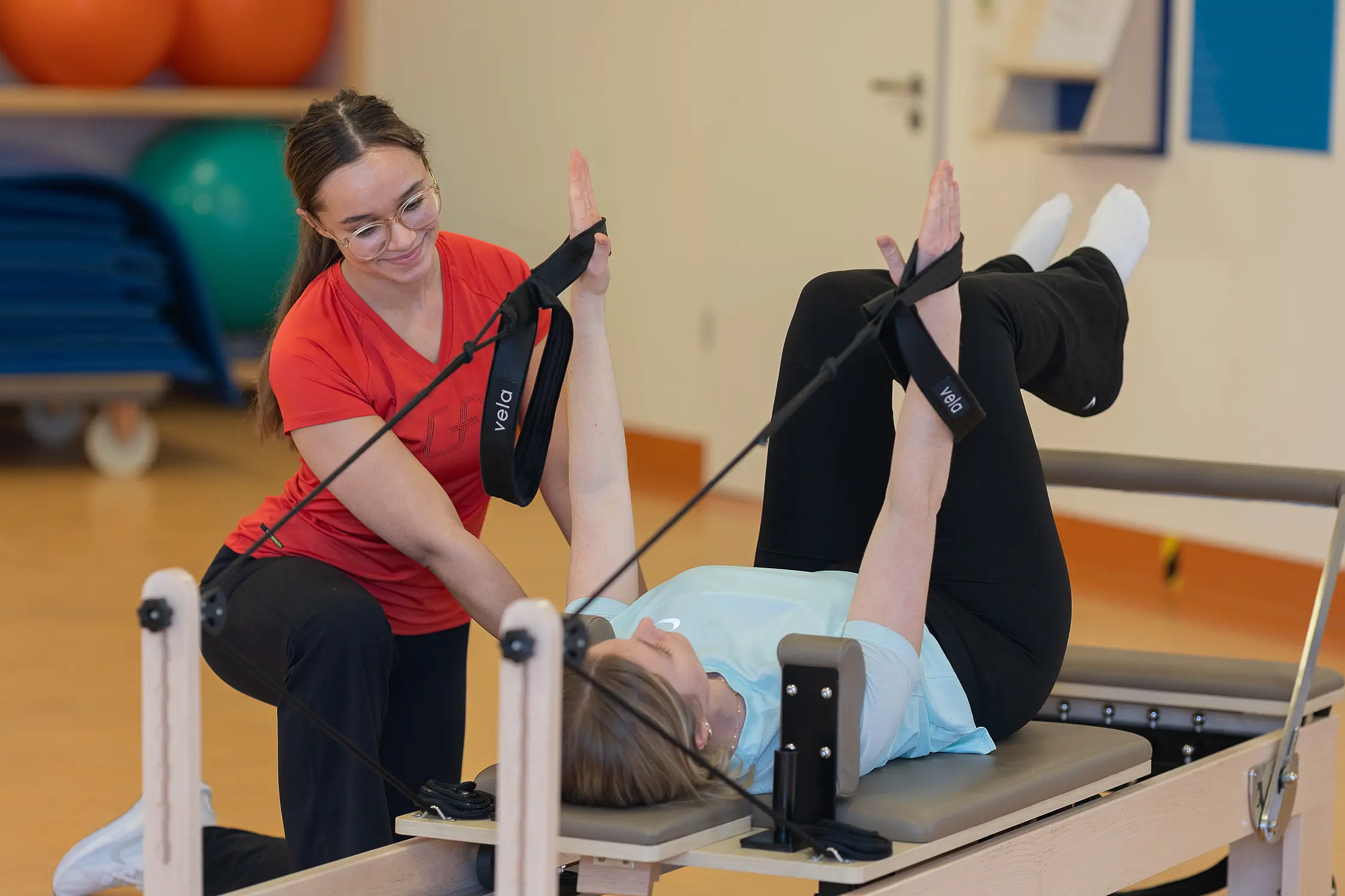 Eine Therapeutin begleitet eine Patientin bei einer Übung auf einem Pilates-Reformer mit Gurten im Therapieraum.