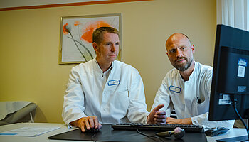 Zwei Ärzte sitzen an einem Schreibtisch und betrachten medizinische Informationen auf einem Computerbildschirm.
