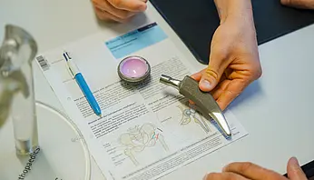 Hände zeigen eine Hüftprothese und erläutern anhand einer Zeichnung, wie das künstliche Hüftgelenk aufgebaut ist und im Körper eingesetzt wird.