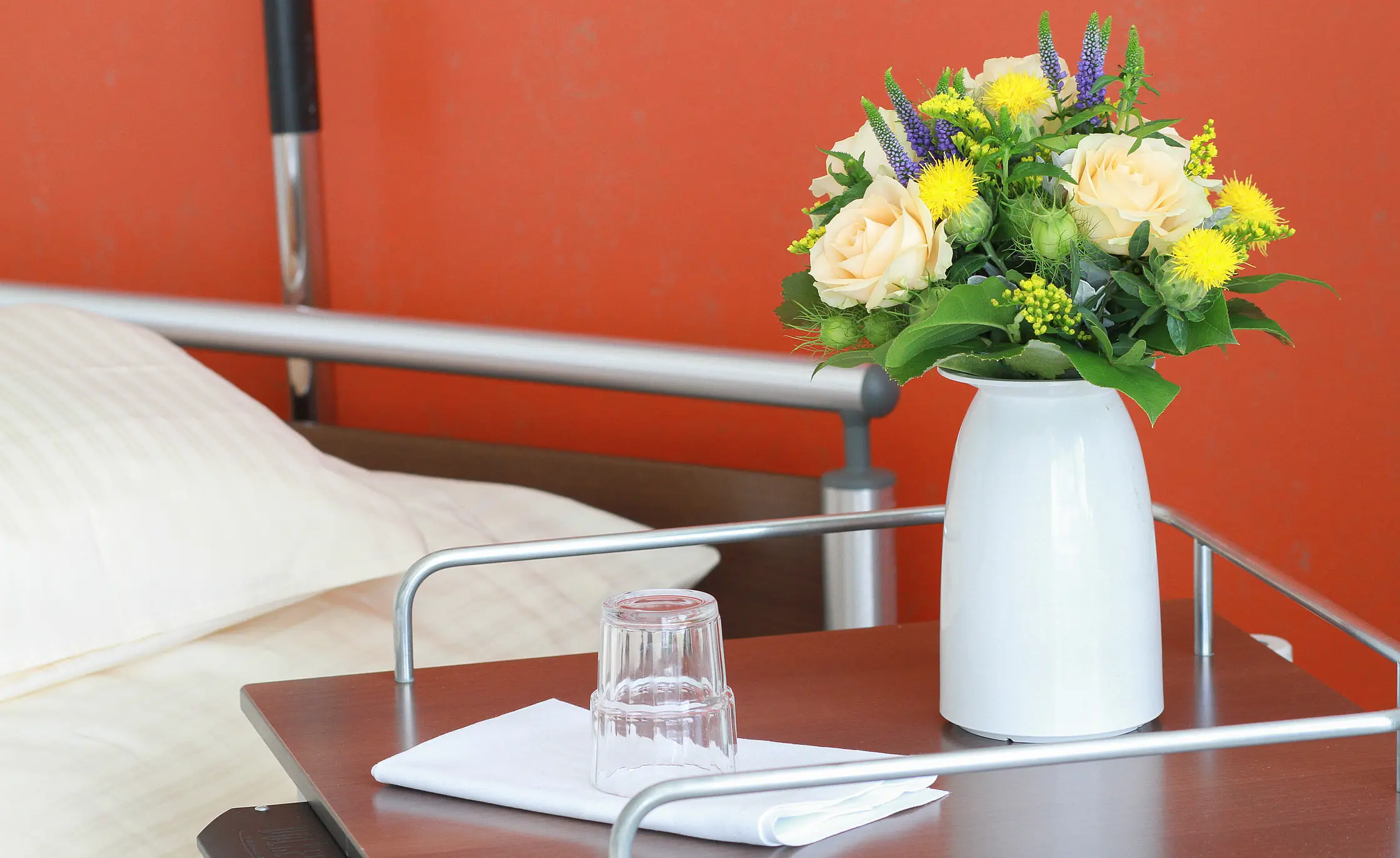 Nachttisch neben einem Pflegebett mit Blumen in einer Vase, Wasserglas und gefaltetem Tuch.