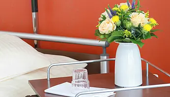 Nachttisch neben einem Pflegebett mit Blumen in einer Vase, Wasserglas und gefaltetem Tuch.