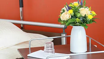 Nachttisch neben einem Pflegebett mit Blumen in einer Vase, Wasserglas und gefaltetem Tuch.