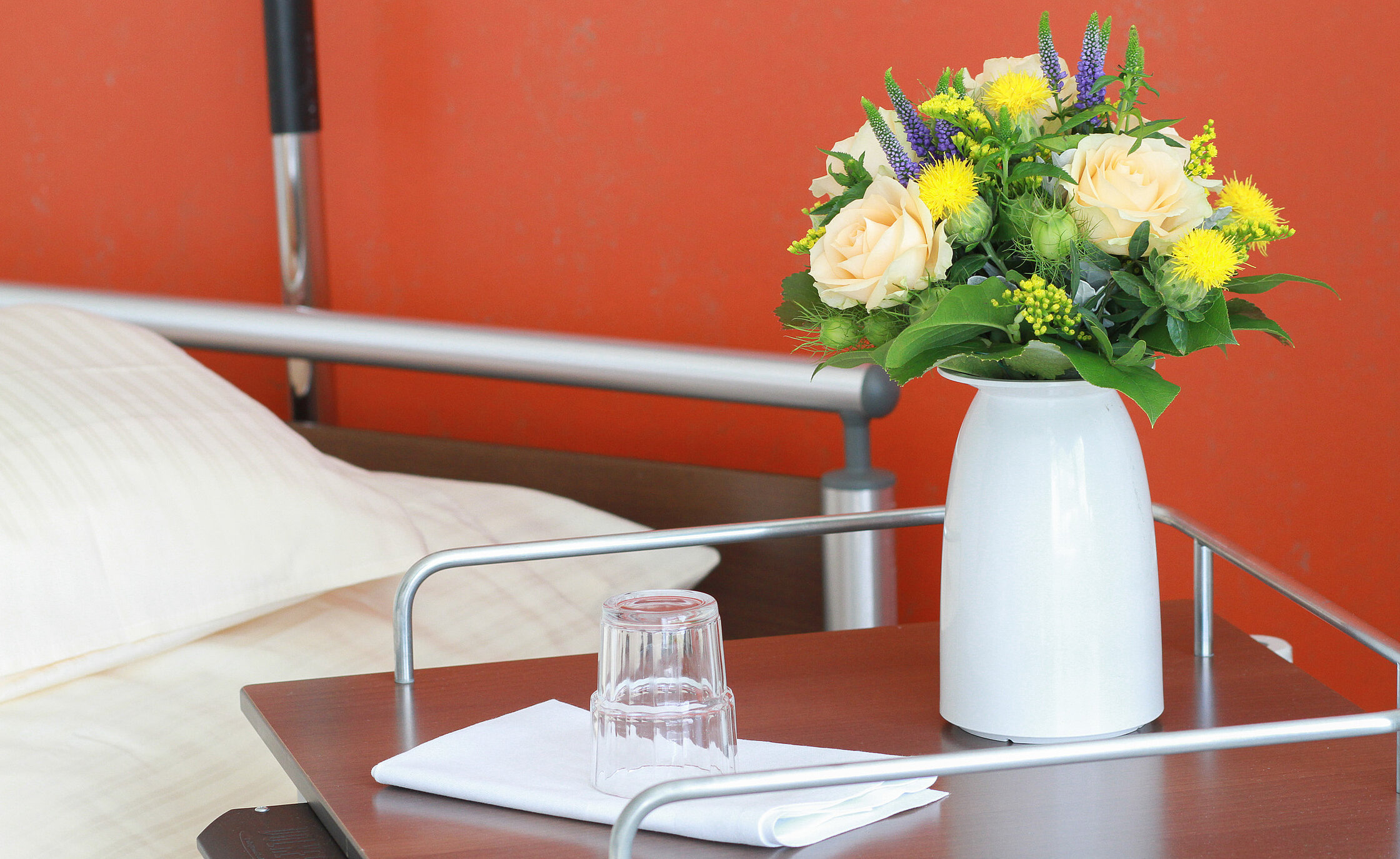 Nachttisch neben einem Pflegebett mit Blumen in einer Vase, Wasserglas und gefaltetem Tuch.