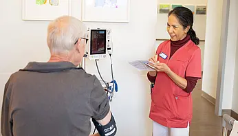 Eine Pflegekraft misst bei einem sitzenden Patienten den Blutdruck und notiert die Werte auf einem Zettel. Beide stehen sich im Patientenzimmer gegenüber.