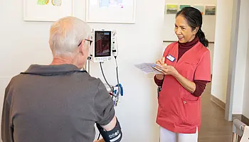 Eine Pflegekraft misst bei einem sitzenden Patienten den Blutdruck und notiert die Werte auf einem Zettel. Beide stehen sich im Patientenzimmer gegenüber.