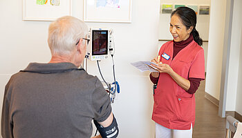 Eine Pflegekraft misst bei einem sitzenden Patienten den Blutdruck und notiert die Werte auf einem Zettel. Beide stehen sich im Patientenzimmer gegenüber.