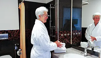 Patientin steht im Bademantel am Waschbecken eines modernen Badezimmers mit Spiegel und Dusche im Hintergrund.