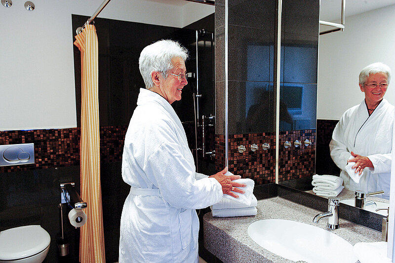 Patientin steht im Bademantel am Waschbecken eines modernen Badezimmers mit Spiegel und Dusche im Hintergrund.
