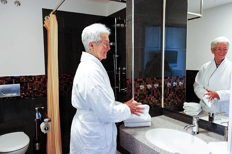 Patientin steht im Bademantel am Waschbecken eines modernen Badezimmers mit Spiegel und Dusche im Hintergrund.