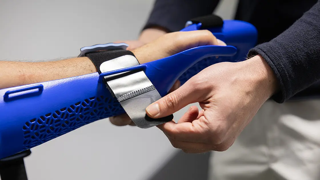 In der orthopädischen Werkstatt stellt ein Orthopädietechniker die Klettverschlüsse einer blauen Handorthese am Unterarm eines Patienten ein.