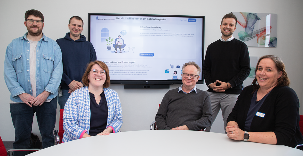 Über das Patientenportal zur Online-Terminvergabe für die Schmerzklinik freut sich die Projektgruppe aus Mitarbeitenden der IT, des Patientenmanagement und der Schmerzklinik. Mitarbeitende sitze am Tisch vor einem Bildschirm, auf dem das Patientenportal zu sehen ist.