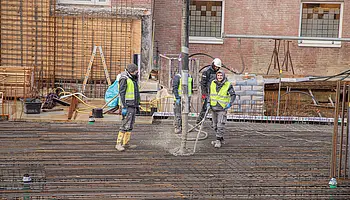 Mehrere Bauarbeitende stehen auf der vorbereiteten Bewehrung und führen den Schlauch der Betonpumpe. Im Hintergrund sind Schalungselemente und das Bestandsgebäude aus Backstein zu sehen.