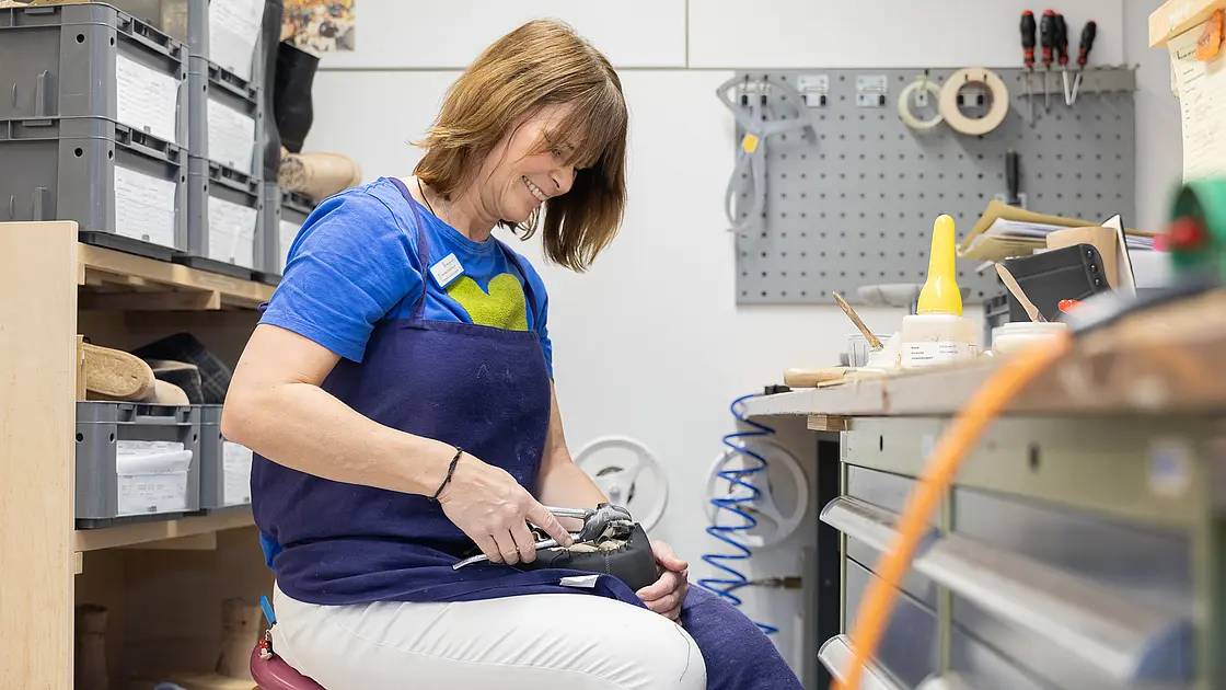 Eine Mitarbeiterin der orthopädischen Werkstatt sitzt an einem Werkbankplatz und bearbeitet einen orthopädischen Schuh mit einem Handwerkzeug; im Hintergrund sind Materialien und Arbeitsgeräte der Schuhtechnik zu sehen.