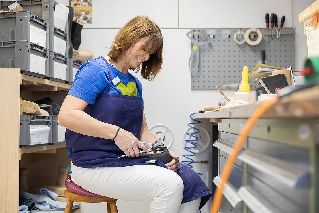 Mitarbeiterin bearbeitet sitzend einen orthopädischen Schuh mit Werkzeug an einem Werkbankplatz.