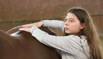 Jugendliche legt beide Arme ruhig auf den Rücken eines Pferdes in einer Halle.