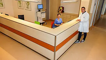 Großer, L-förmiger Tresen mit Mitarbeitenden in Dienst- und Arztkleidung in einem offenen Stationsbereich.