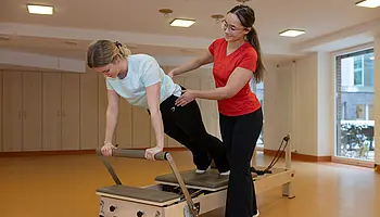 Eine Patientin führt eine Übung im Vierfüßstand auf einem Pilates-Reformer aus, begleitet von einer Therapeutin.