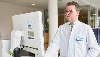 Ein Arzt im weißen Kittel steht an einer mobilen Monitorstation und betrachtet medizinische Bilddaten im Klinikflur.